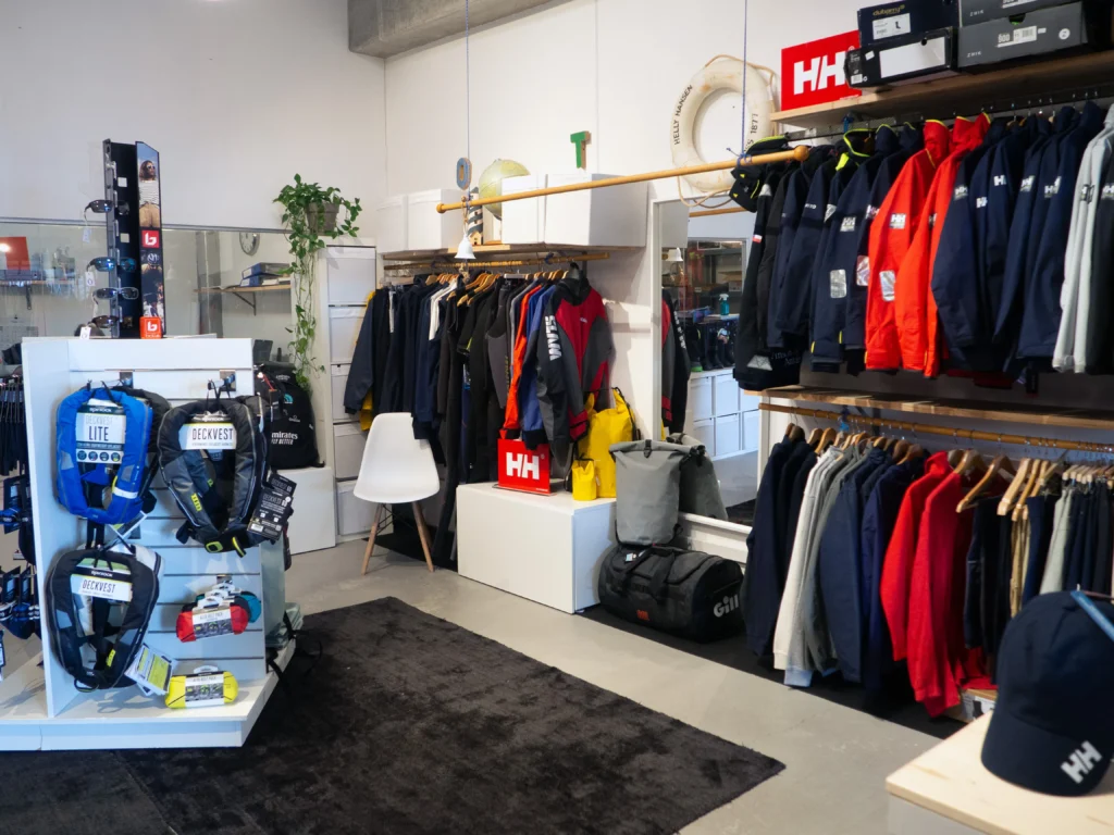 Interior de una tienda náutica con una amplia selección de chaquetas técnicas Helly Hansen, un maniquí con traje de supervivencia rojo y un mostrador central con vitrinas de dispositivos Garmin
