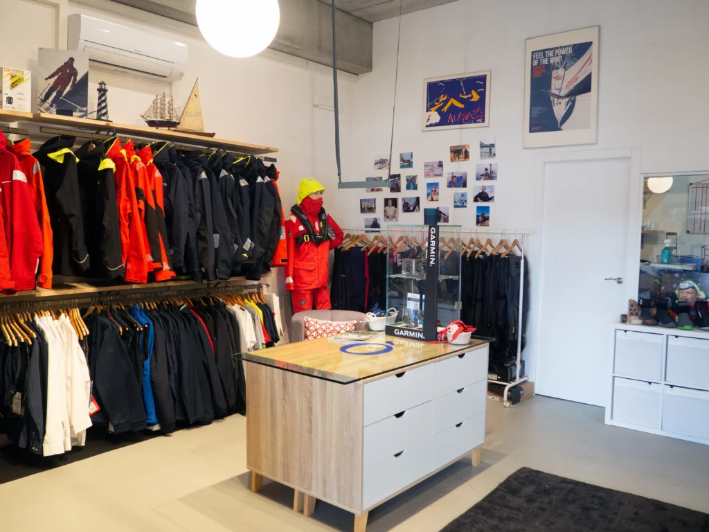 Interior de una tienda náutica con una amplia selección de chaquetas técnicas Helly Hansen, un maniquí con traje de supervivencia rojo y un mostrador central con vitrinas de dispositivos Garmin.
