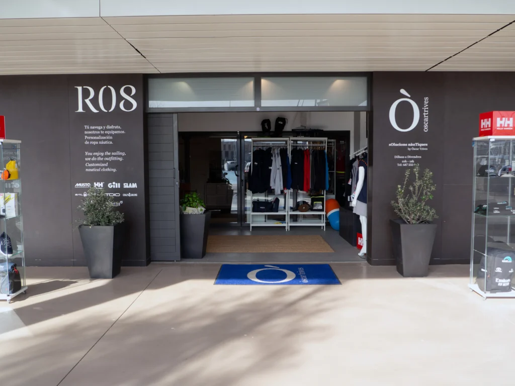 Fachada exterior de la tienda 'Ò oscartrives sOlucions nàuTiques', mostrando la entrada principal con logotipos de marcas como Musto, Helly Hansen y Slam, y un felpudo azul corporativo.