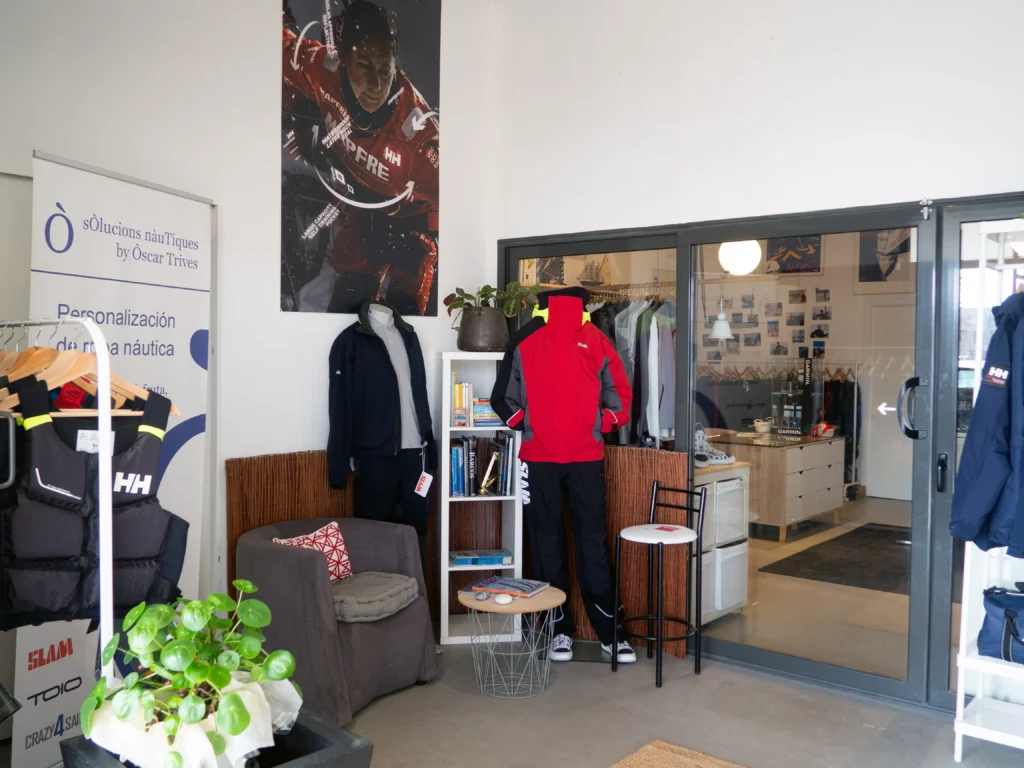 Zona de bienvenida en la tienda náutica de Óscar Trives, con un sillón de descanso, maniquíes con chaquetas técnicas de Helly Hansen y Slam, y cartelería sobre personalización de ropa náutica.