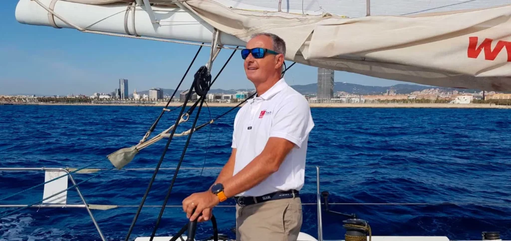 Hombre con polo blanco y gafas de sol manejando el timón de un barco de vela en aguas del Mediterráneo, con una vista panorámica de Barcelona y sus edificios emblemáticos detrás.